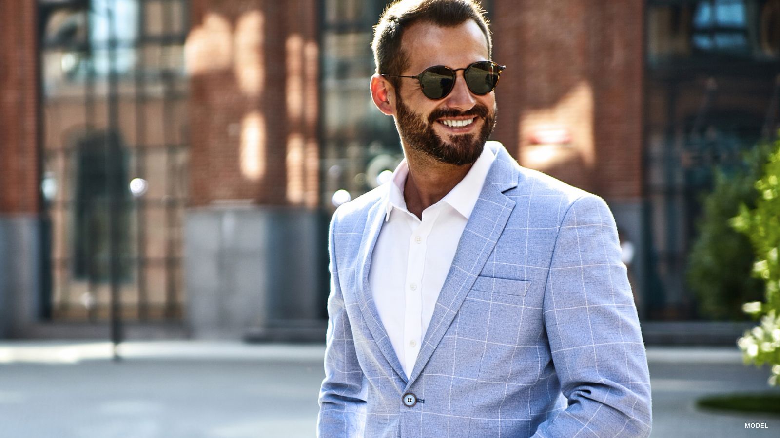 Man wearing sunglasses standing outside smiling, wearing a white button up shirt and a light blue jacket